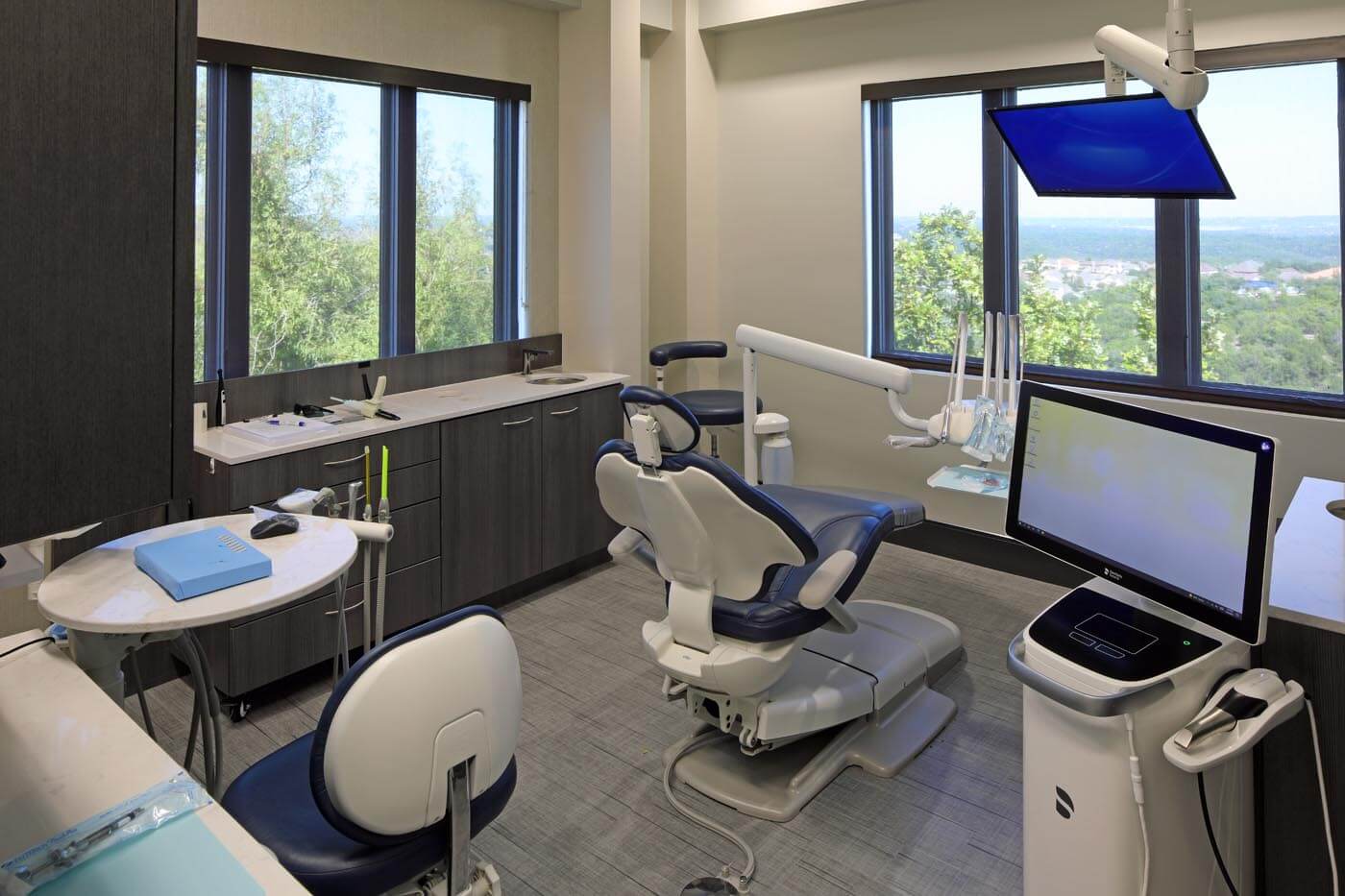 A view inside one of SkyRidge Dental's clean and sleek exam rooms, where various technology alongside an exam chair can be seen. A beautiful view of Lakeway, Texas can be seen outside the large windows.