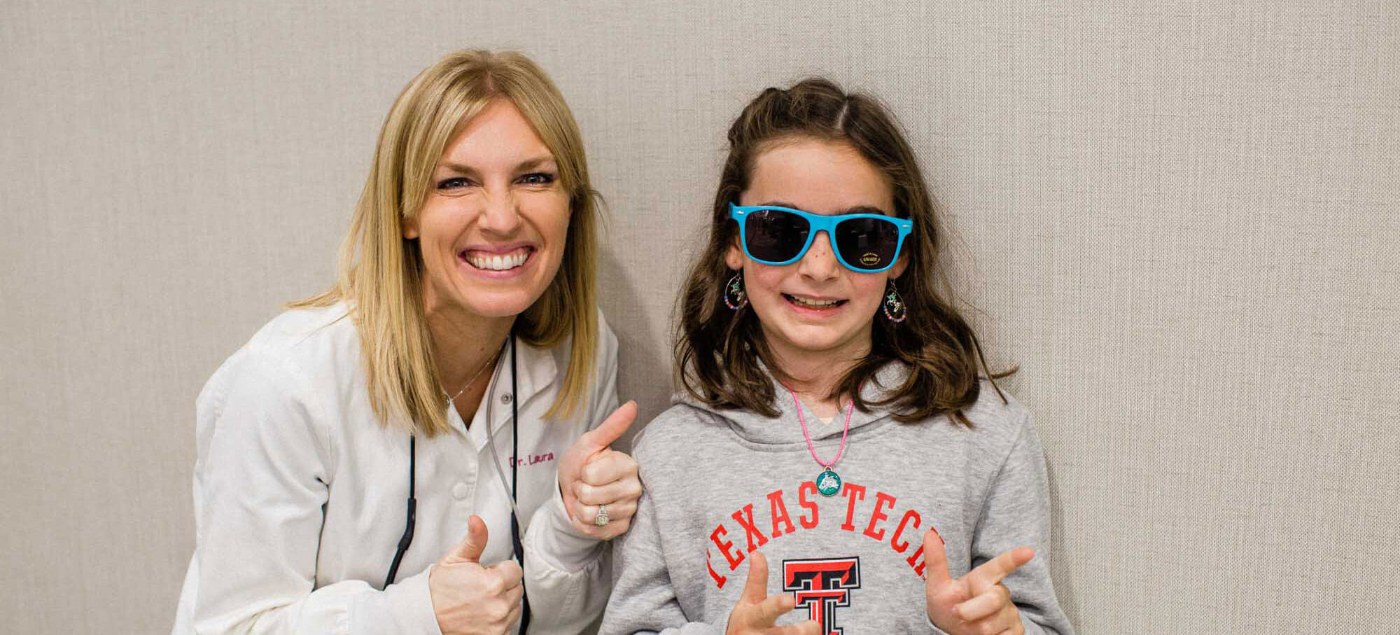 Dr. Cook smiles and gives a thumbs-up with a young patient, who wears protective dental exam sunglasses and smiles while giving two playful finger-guns.